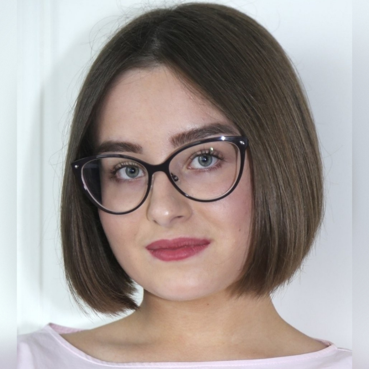 A woman with bobbed brown hair wearing cat-eye glasses and a pink top. She has blue eyes and rose-pink lipstick.